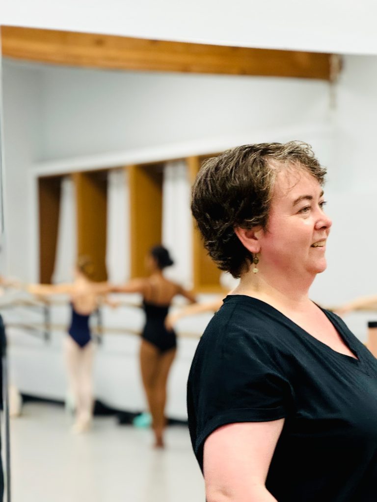 Ballet teacher standing and smiling during class with dancers reflected in the mirror behind her